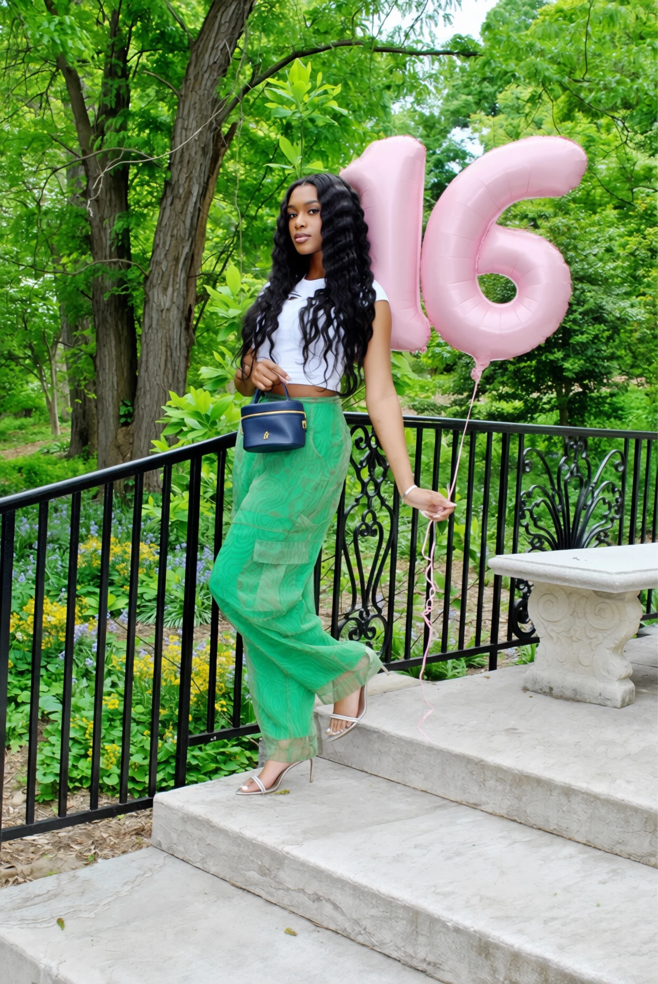 Sweet 16 birthday portrait on garden steps, with pink number 16 balloons, a white top, green trousers, and a blue handbag