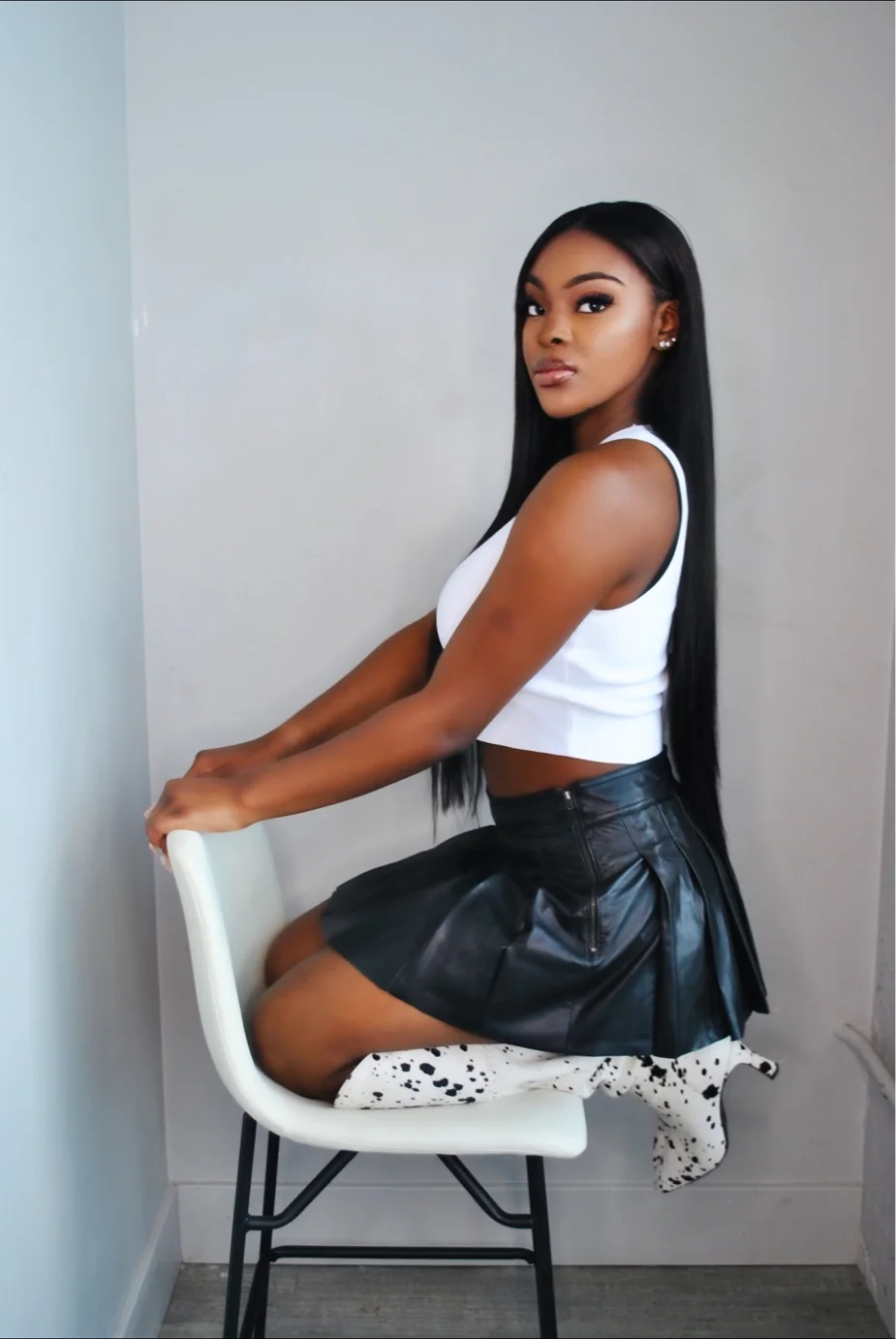 Model in a white tank top and black pleated skirt kneeling backward on a white chair, studio editorial